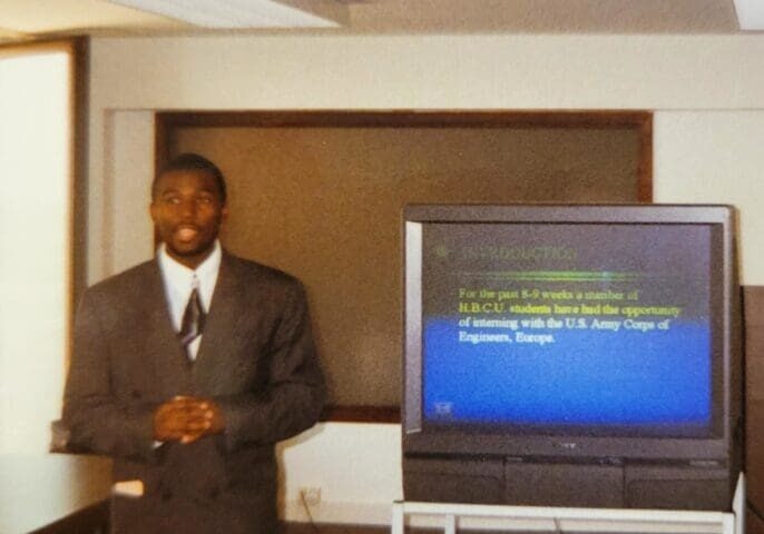 Man in suit presenting beside television monitor